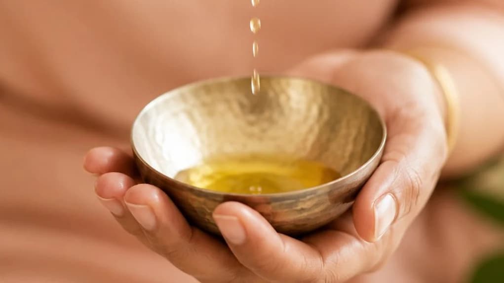 Antique copper bowl of golden Ayurvedic oil with fresh tulsi, neem, and turmeric on warm linen — Ayurveda at The Wellness Ed.