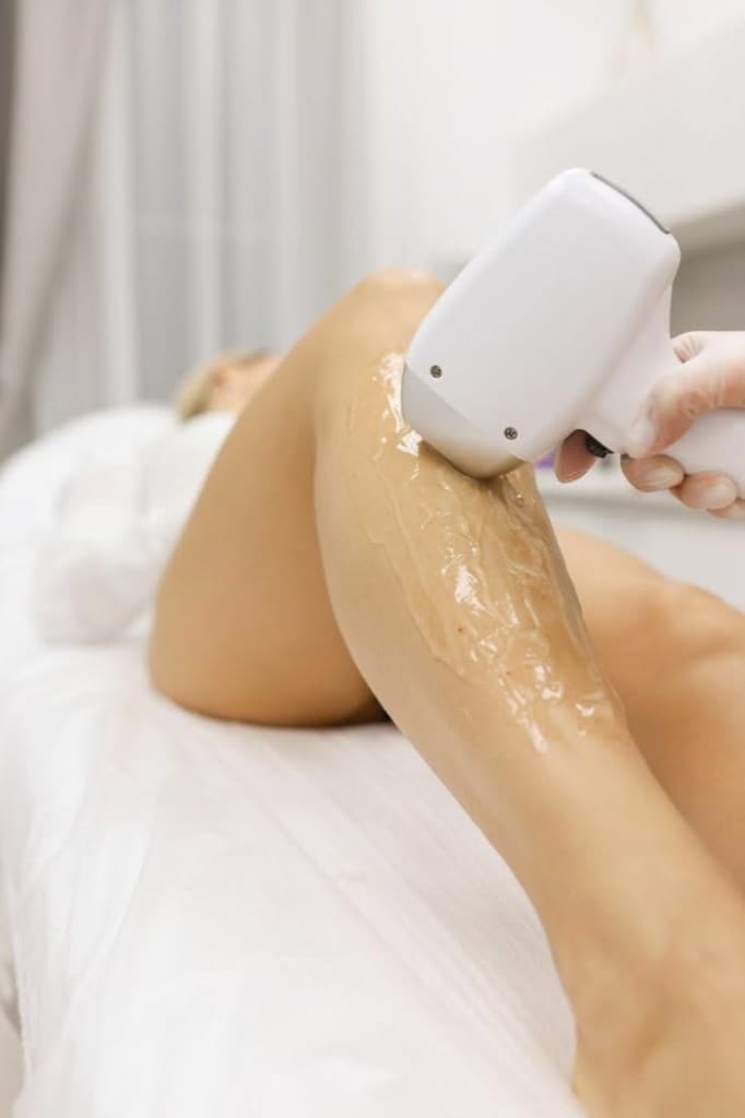 Practitioner gliding a modern laser handpiece across a guest's leg coated in cooling clear gel, in a softly-lit treatment room — Laser care at The Wellness Ed.