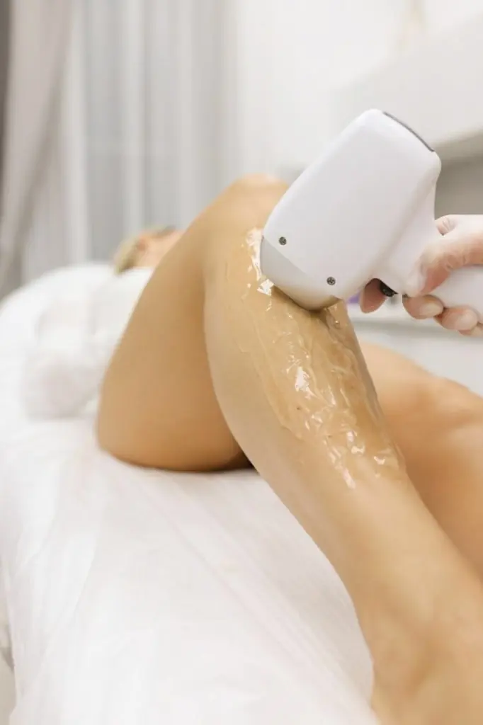 Practitioner gliding a modern laser handpiece across a guest's leg coated in cooling clear gel, in a softly-lit treatment room — Laser care at The Wellness Ed.