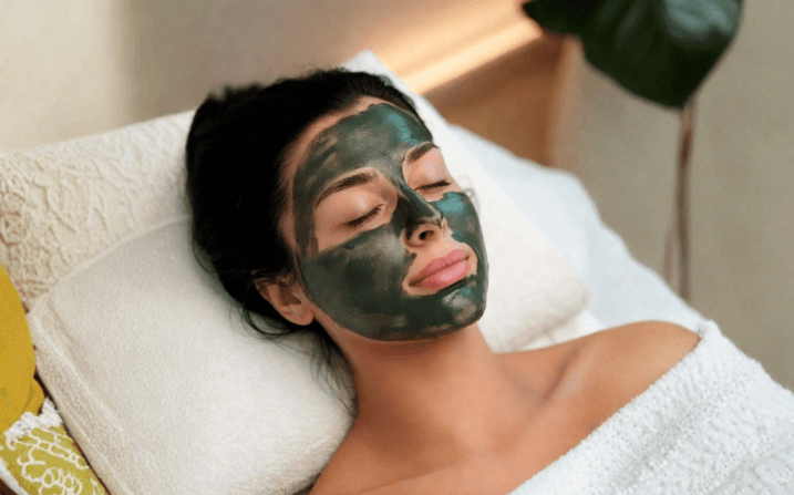 A guest unwinding during an Elixir Pro facial at The Wellness Ed — a calming, deeply hydrating green botanical mask resting on her skin while she relaxes against a soft towel, a sculptural monstera leaf framing the warm-lit treatment room.
