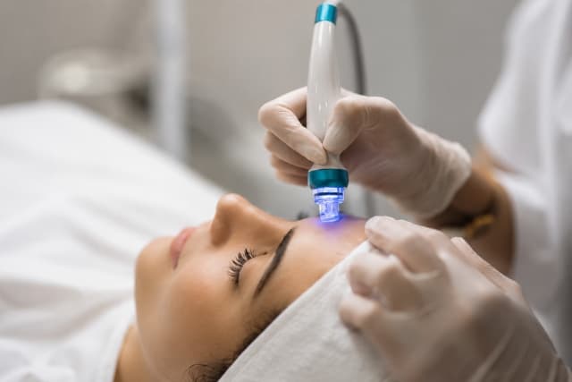 Close-up of a Hydrafacial session at The Wellness Ed — a gloved clinician glides the signature blue-lit vortex handpiece across a relaxed guest's forehead, cleansing, exfoliating and hydrating the skin in one calm, restorative pass.