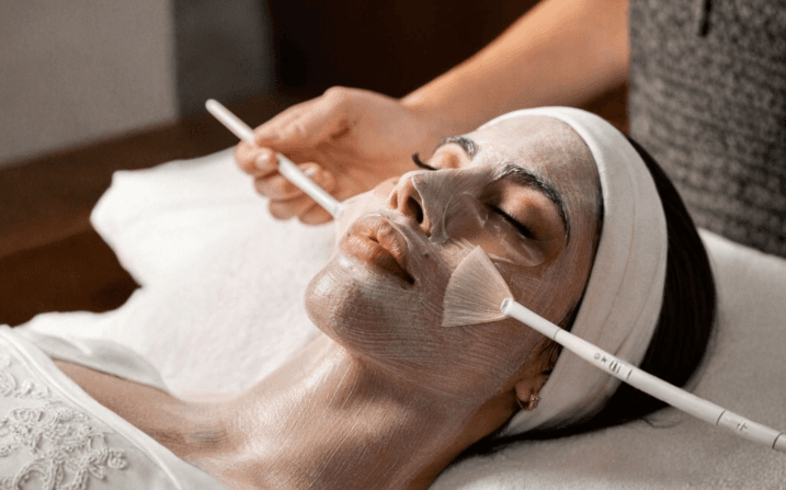 A guest mid-treatment during a Kempeki Facial at The Wellness Ed — a therapist's fan brush sweeping a luminous silver-clay mask across the skin, the soft headband and warm-lit room reflecting the ritual's calm, precise, Japanese-inspired cadence.