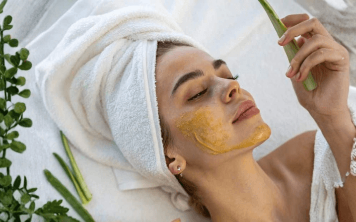A guest enjoying the Wellness Ed Signature Facial — golden honey-and-fresh-fruit mask warming her skin while she reclines in a fluffy white towel turban, a fresh aloe-vera leaf resting in her hand and lush green foliage framing the sunlit treatment room at The Wellness Ed.