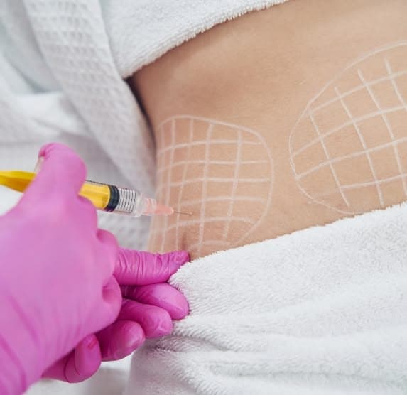 Close-up of a Fat X session at The Wellness Ed — a clinician in pink gloves administers a precise fat-dissolving micro-injection into a mapped grid on a guest's abdomen, with a soft white towel drape and clinical-clean styling.