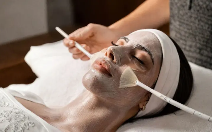 A guest mid-treatment during a Kempeki Facial at The Wellness Ed — a therapist's fan brush sweeping a luminous silver-clay mask across the skin, the soft headband and warm-lit room reflecting the ritual's calm, precise, Japanese-inspired cadence.