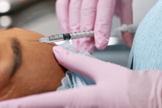 Close-up of a Skin Boosters session at The Wellness Ed — a clinician in pink medical gloves precisely administers a fine micro-injection of skin-quality hydrators into a guest's forehead, the steady, gentle technique characteristic of an in-clinic, doctor-led protocol.
