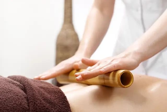 A guest enjoying a Bamboo Therapy session at The Wellness Ed — a therapist in white linen rolls a smooth, polished bamboo cylinder along the contours of the back with gentle, even pressure, the warm-toned treatment room and soft brown towel reflecting the ritual's calm, grounding cadence.