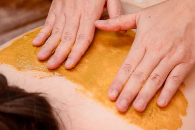 A guest receiving a warming Herbal Body Wrap at The Wellness Ed — a therapist's hands press a rich, earthy herbal paste evenly across the back, surrounded by fresh green leaves and small bowls of botanicals.