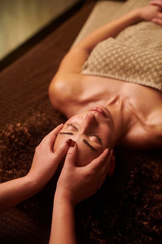 A guest receiving an Organic Facial at The Wellness Ed — gentle hands cradle the jaw in slow, rhythmic strokes during a warm, dimly lit treatment that completes the body-care ritual with a moment of stillness.