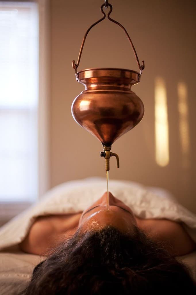 A guest receiving Shirodhara at The Wellness Ed — a polished copper vessel suspended above her releases a continuous, golden thread of warm herbal oil onto the centre of her forehead, the warm-lit treatment room and white linen draping setting a deeply meditative, restorative tone.