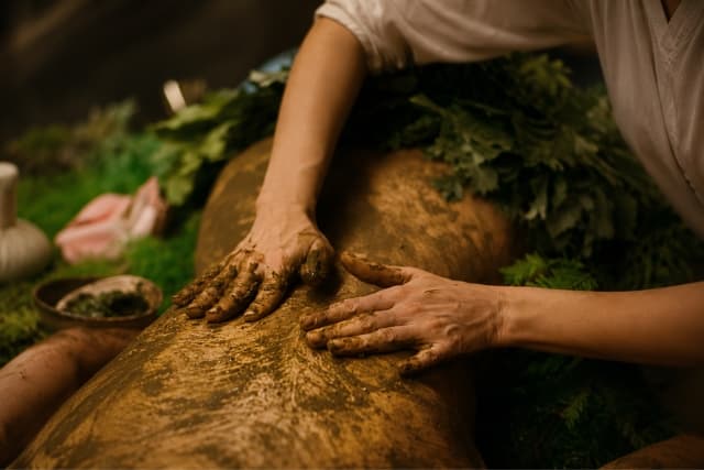 A therapist's hands smoothing a warm, golden herbal powder paste in long, rhythmic strokes across a guest's back during an Udwarthanam dry-powder massage at The Wellness Ed.